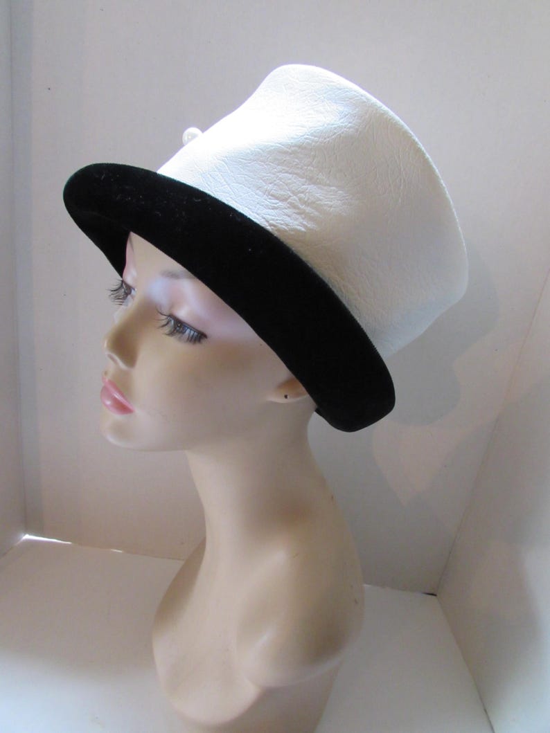 May include: A white hat with a black brim. The hat has a slightly curved top and a black band around the base. A small, white pearl-like embellishment is visible on the side. The hat is displayed on a mannequin head.