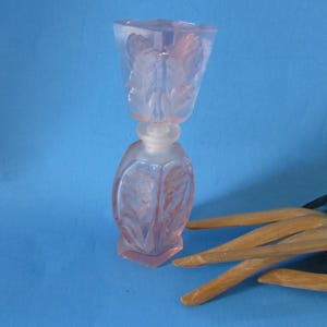 May include: A pink glass perfume bottle with a geometric design and a square stopper. The bottle has a frosted glass stopper and is decorated with leaf-like patterns. The bottle is on a blue background.