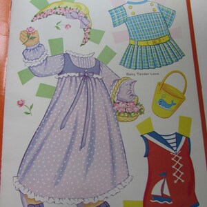 May include: A page from a vintage paper doll book featuring several outfits. The outfits include a long lavender dress with white polka dots, a blue and white plaid dress, and a red sailor-style romper with a sailboat graphic.