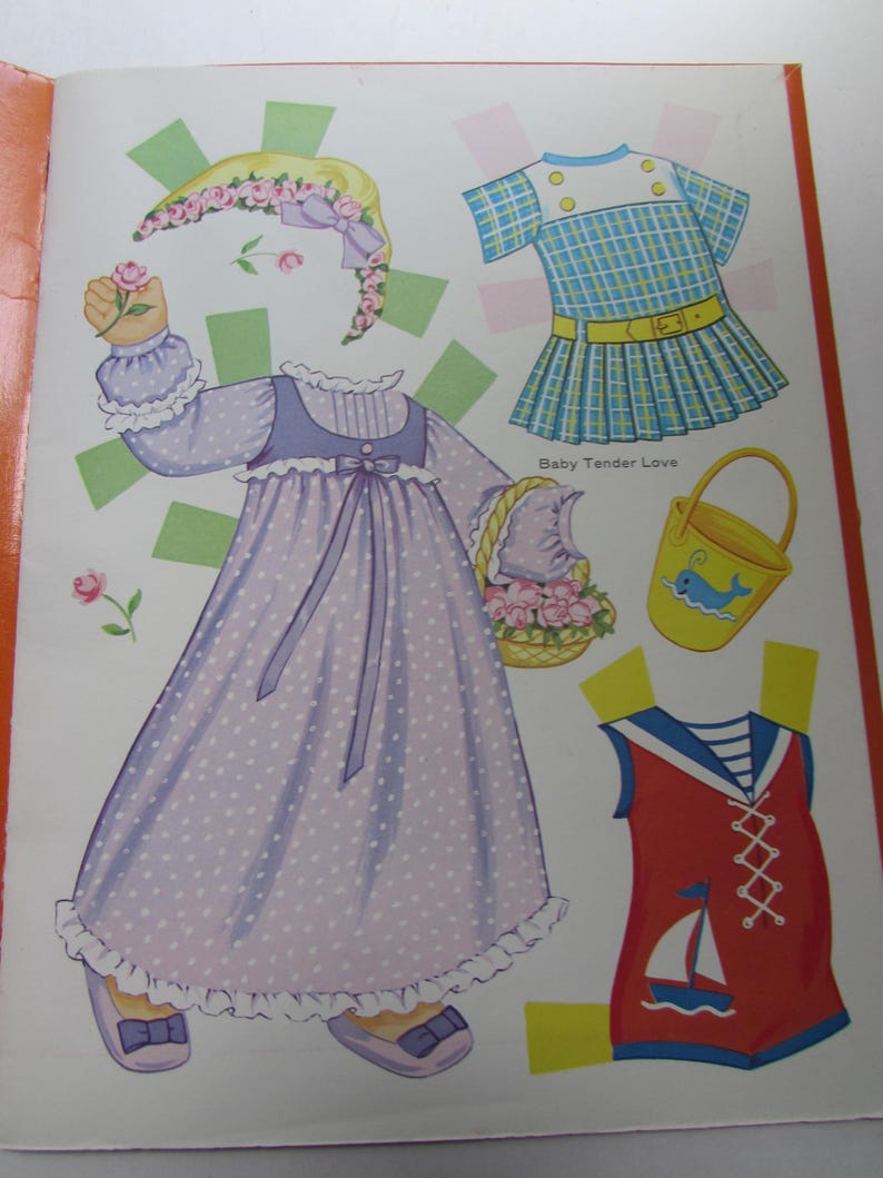 May include: A page from a vintage paper doll book featuring several outfits. The outfits include a long lavender dress with white polka dots, a blue and white plaid dress, and a red sailor-style romper with a sailboat graphic.