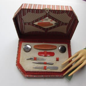 Puede incluir: Un estuche de tocador vintage de rayas rojas y blancas con un espejo y un juego de herramientas de manicura, que incluyen una lima de uñas, unas pinzas y un empujador de cutículas.