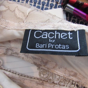 May include: A black and white label with the text "Cachet by Bari Protas" on a white lace fabric.
