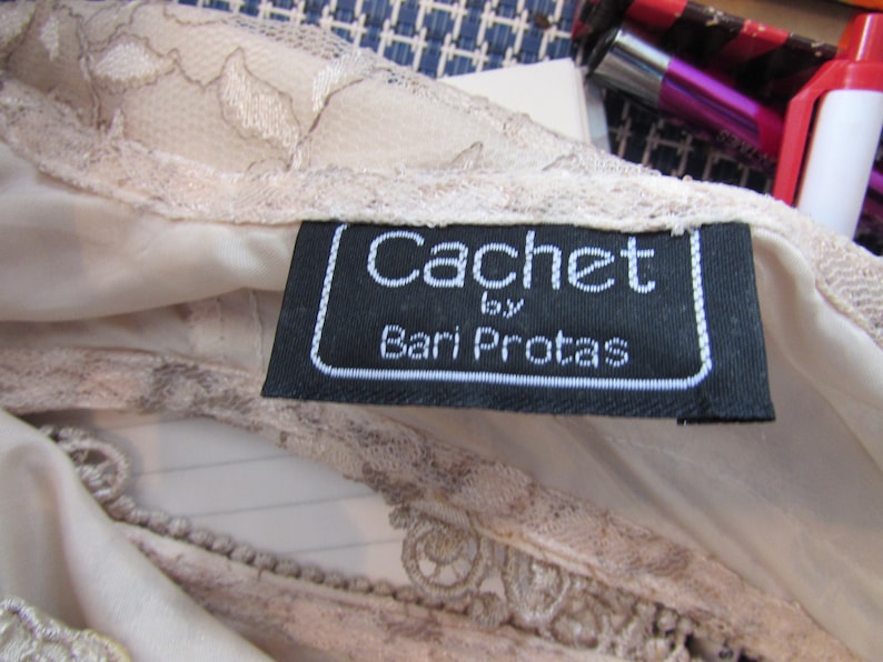 May include: A black and white label with the text "Cachet by Bari Protas" on a white lace fabric.