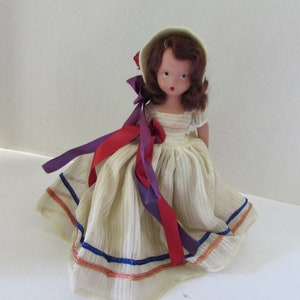 May include: A vintage doll with brown hair, wearing a white dress with blue and orange trim, and a white bonnet with red and purple ribbons.