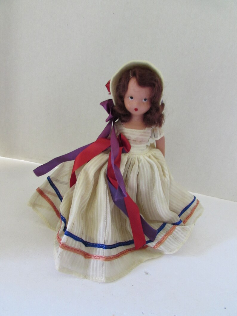 May include: A vintage doll with brown hair, wearing a white dress with blue and orange trim, and a white bonnet with red and purple ribbons.
