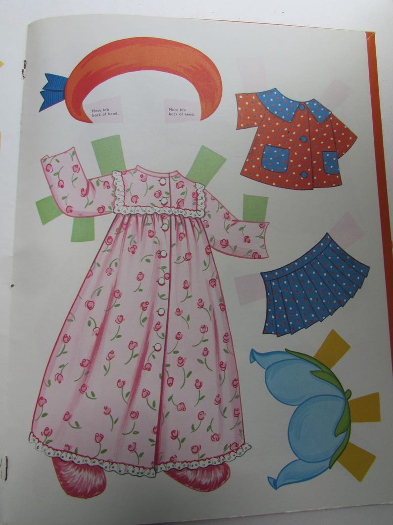 May include: A vintage paper doll page featuring cut-out clothing and accessories. The clothing includes a pink floral nightgown, a red polka-dot top, a blue skirt with white dots, and a blue flower. An orange hat with a blue bow is also included.