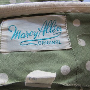 May include: A white label with blue embroidered text that reads "Marcy Allen Original" on a green fabric with white polka dots.