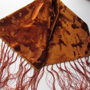 May include: A brown scarf with a textured pattern and long, reddish-brown fringe. The scarf has a soft, velvety appearance and is folded over, showcasing its design. The fringe hangs from the edges.