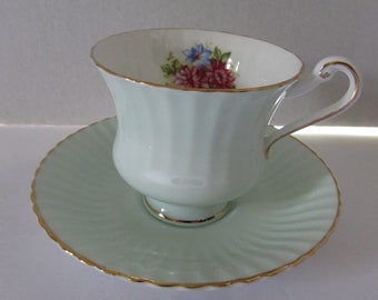 May include: A light green teacup and saucer set with a floral design. The cup has a delicate handle and a gold rim. The saucer has a scalloped edge and a gold rim.