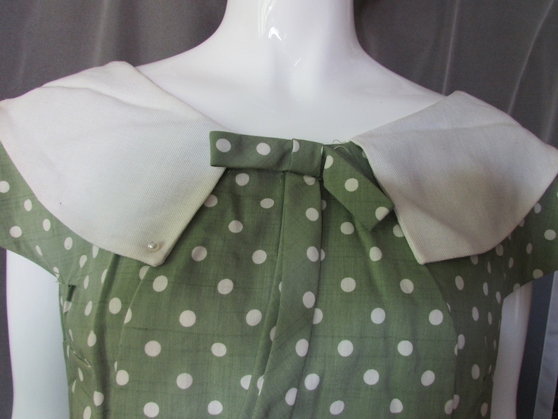 May include: A green dress with white polka dots and a white collar. The dress has a bow at the neckline.