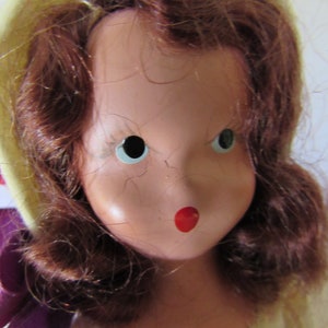 May include: A vintage porcelain doll with red and white painted lips, blue eyes, and brown hair styled in a curly bob. The doll is wearing a yellow hat and a purple ribbon.