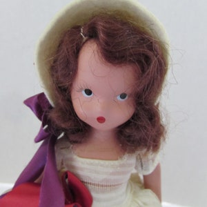May include: A vintage porcelain doll with brown curly hair, wearing a white dress and a white hat with a purple ribbon. The doll has painted features, including red lips and blue eyes.