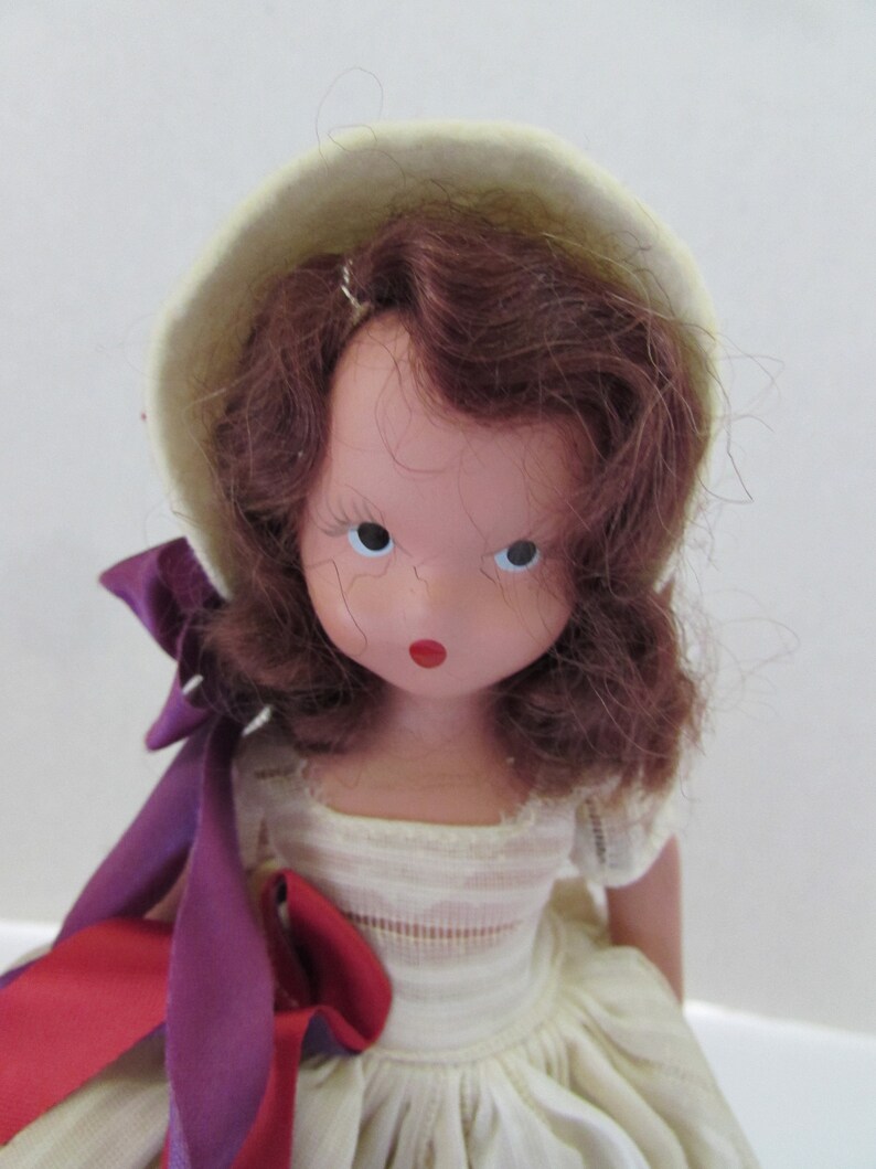 May include: A vintage porcelain doll with brown curly hair, wearing a white dress and a white hat with a purple ribbon. The doll has painted features, including red lips and blue eyes.