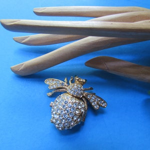 May include: A gold bee brooch with clear rhinestones. The bee is facing right and has its wings spread out. The brooch is on a blue background.