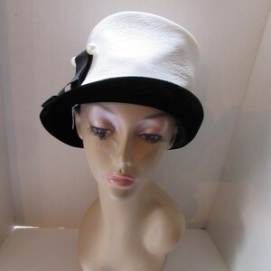 May include: A white hat with a textured surface and a black velvet brim. A black ribbon and a pearl accent the side of the hat. The hat is displayed on a mannequin head.