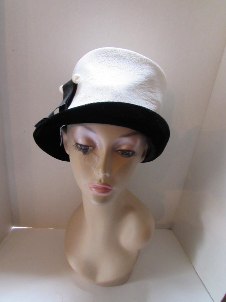 May include: A white hat with a textured surface and a black velvet brim. A black ribbon and a pearl accent the side of the hat. The hat is displayed on a mannequin head.