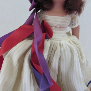 May include: A vintage doll with brown hair wearing a white dress with a ruffled skirt and a purple and red ribbon tied around her waist.