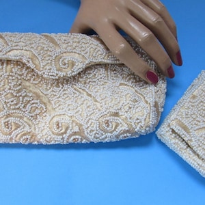 May include: A set of two vintage beaded purses. The larger purse is a clutch with a flap closure. The smaller purse is a compact. Both purses are made of a cream-colored fabric covered in white beads.