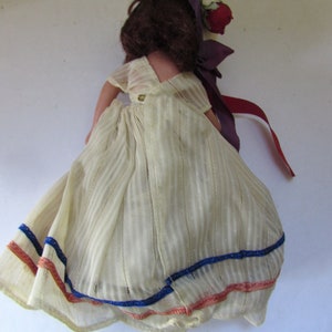 May include: A vintage doll with brown hair, wearing a white dress with blue and red trim. The doll is wearing a purple hat with a red flower.