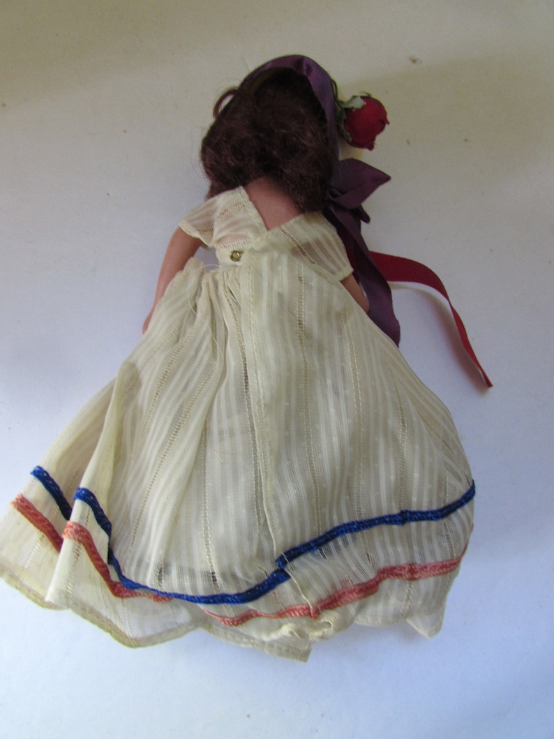 May include: A vintage doll with brown hair, wearing a white dress with blue and red trim. The doll is wearing a purple hat with a red flower.
