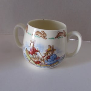 May include: Cream-colored ceramic mug with two handles, featuring a charming illustration of rabbits in a boat. The design includes rabbits in red and blue clothing, with additional running rabbit motifs around the rim. A classic children's design.