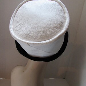 May include: A white hat with a circular top and a black band. The hat has a textured surface and a rounded brim. The hat is displayed on a mannequin head.