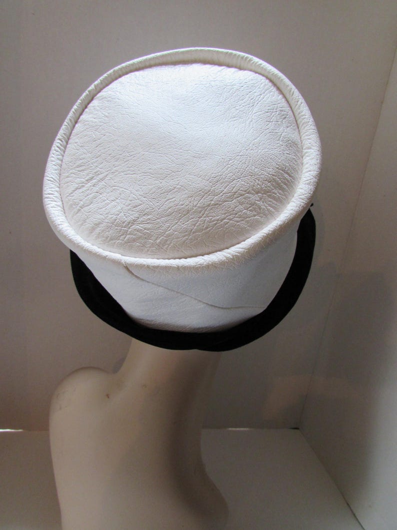 May include: A white hat with a circular top and a black band. The hat has a textured surface and a rounded brim. The hat is displayed on a mannequin head.