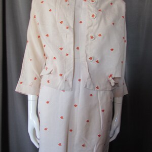 May include: A white dress and jacket set with a small orange floral pattern. The jacket has a button closure and pockets. The dress has a simple, straight silhouette.