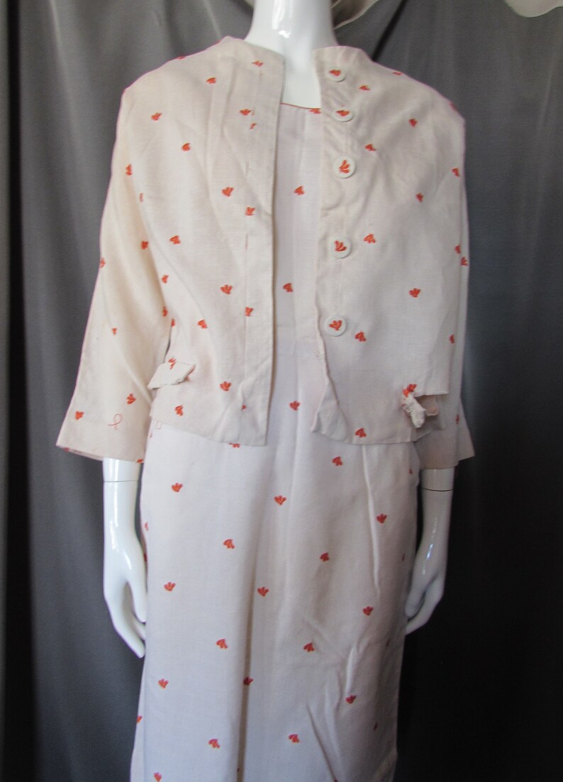 May include: A white dress and jacket set with a small orange floral pattern. The jacket has a button closure and pockets. The dress has a simple, straight silhouette.