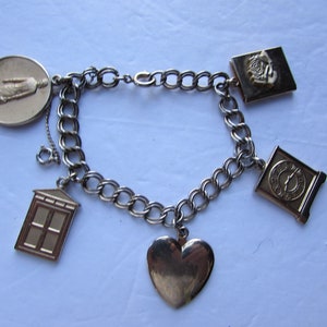 May include: A silver charm bracelet with five charms: a heart, a clock, a house, a figure, and a book. The charms are attached to a silver chain.