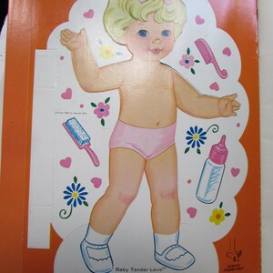 May include: A vintage paper doll cutout of a baby with blonde hair and a pink bow. The baby is wearing pink underwear, white socks, and blue shoes. The image includes the text "Baby Tender Love".