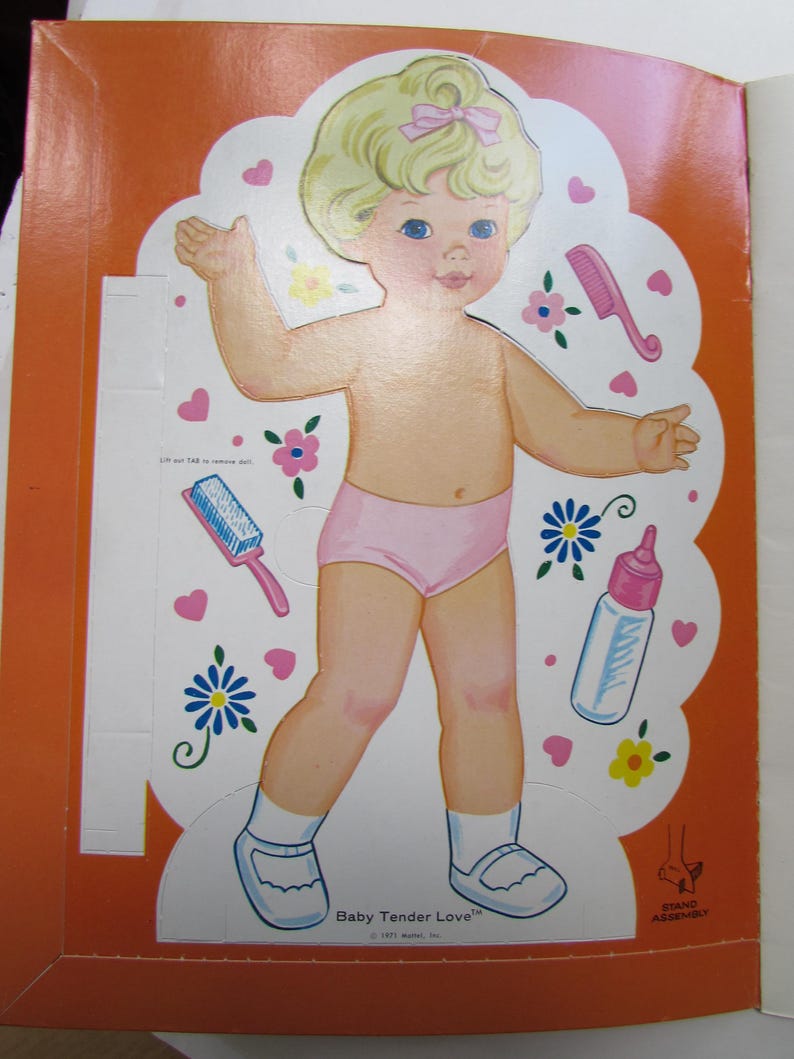 May include: A vintage paper doll cutout of a baby with blonde hair and a pink bow. The baby is wearing pink underwear, white socks, and blue shoes. The image includes the text "Baby Tender Love".