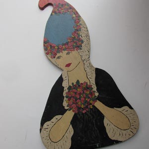 May include: Wooden folk art figure of a woman in a black dress holding a bouquet of flowers. The figure has a blue wig with pink and red flowers, and a red lip. The figure is painted on wood.