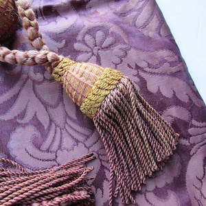 May include: A close-up of a purple fabric with a floral pattern. A brown and gold tassel hangs from the fabric. The tassel has a braided cord and a fringe.