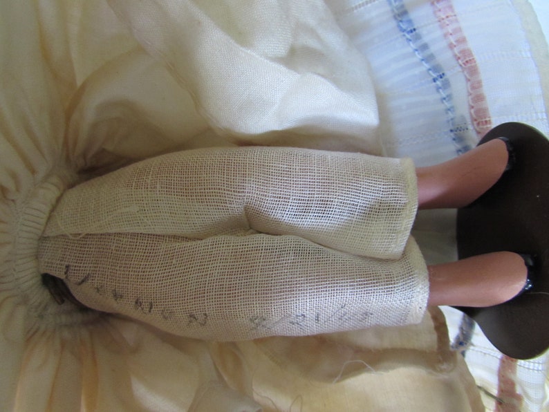 May include: Close-up of a doll's legs wearing white fabric pants with a mesh texture. The fabric has the words "Queen" and "Princess" written on it. The doll's legs are made of porcelain and are wearing black shoes.