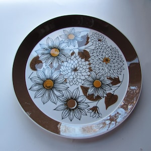 Könnte beinhalten: Ein brauner und weißer Keramikteller mit Blumenmuster. Der Teller hat einen braunen Rand und eine weiße Mitte mit einem Muster aus stilisierten Blumen in Braun-, Weiß- und Gelbtönen.