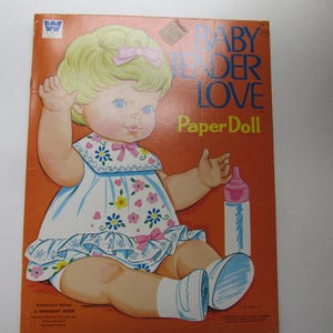 May include: Vintage "Baby Tender Love Paper Doll" book cover. The cover features a cartoon baby doll with blonde hair, a pink bow, and a white dress with blue and pink floral accents. A baby bottle is next to the doll on an orange background.