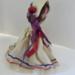 May include: A vintage doll with a white dress, red and purple ribbons, and a white bonnet with a red rose. The doll has brown hair and painted eyes.