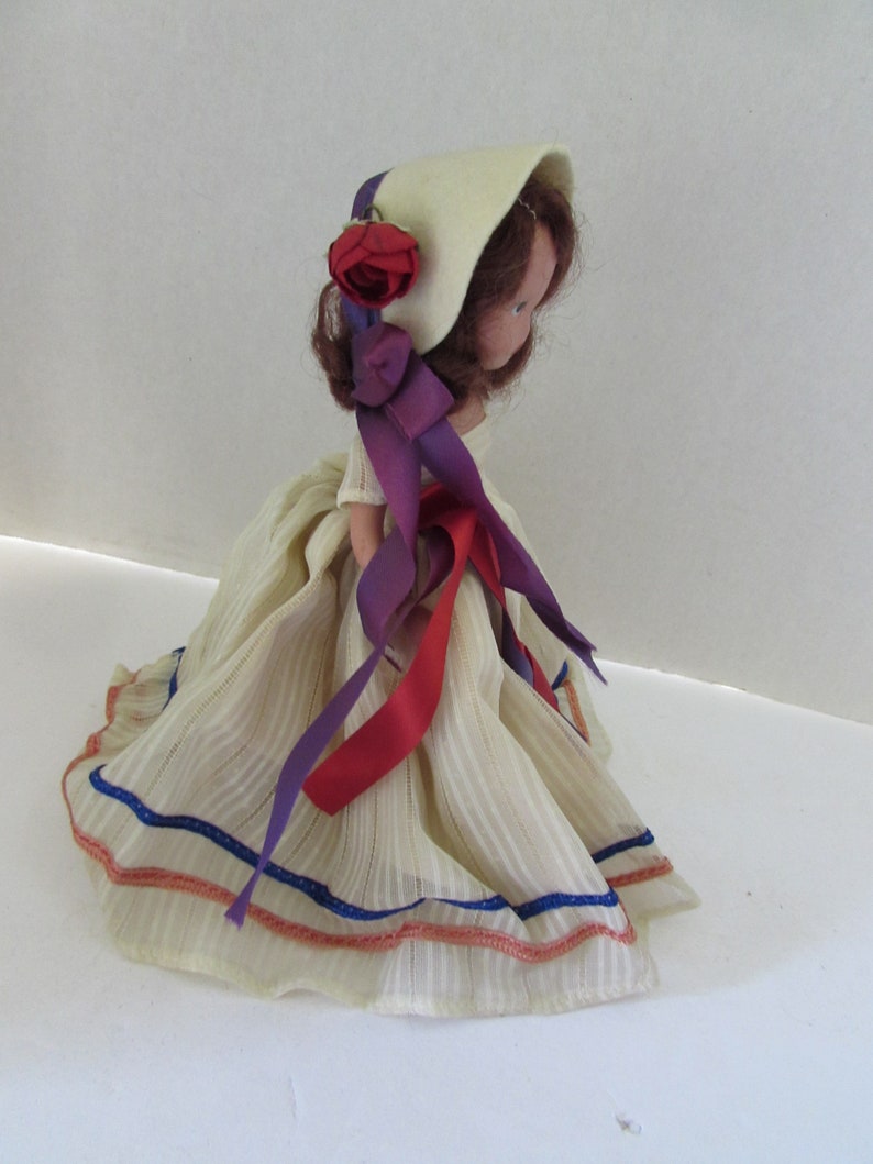 May include: A vintage doll with a white dress, red and purple ribbons, and a white bonnet with a red rose. The doll has brown hair and painted eyes.
