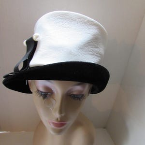 May include: A vintage white hat with a textured surface and a black velvet brim. The hat features a black ribbon and a pearl-like accent. This classic hat is a stylish accessory.