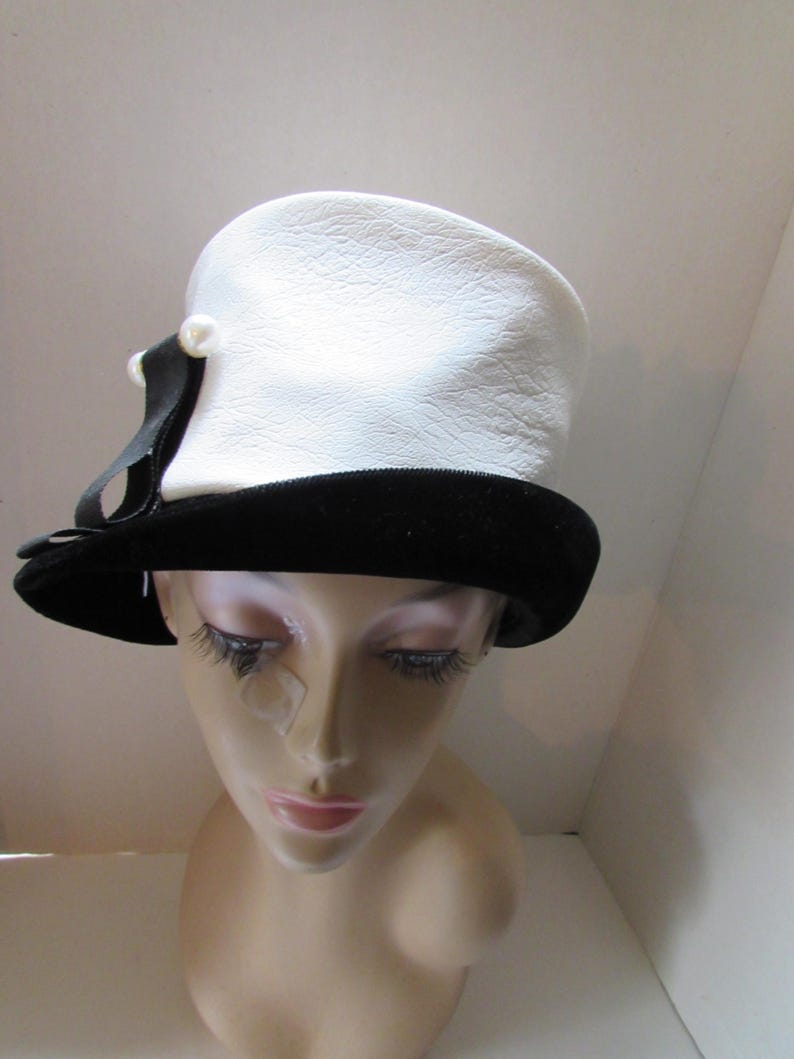 May include: A vintage white hat with a textured surface and a black velvet brim. The hat features a black ribbon and a pearl-like accent. This classic hat is a stylish accessory.