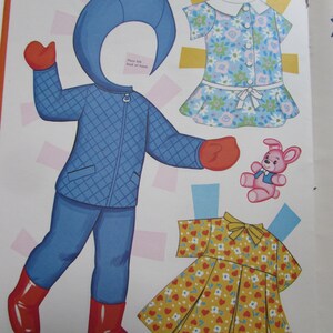 May include: A vintage paper doll set with a blue quilted winter outfit, red boots and mittens, and two dresses. One dress is floral, and the other is yellow with red hearts. A pink teddy bear is also included.