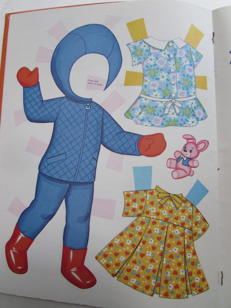 May include: A vintage paper doll set with a blue quilted winter outfit, red boots and mittens, and two dresses. One dress is floral, and the other is yellow with red hearts. A pink teddy bear is also included.