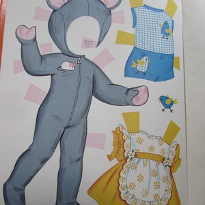 May include: A vintage paper doll set featuring a gray mouse costume with pink accents, a blue and white checkered outfit with bird graphics, and a yellow dress with a white floral apron. The set is open on a white background.