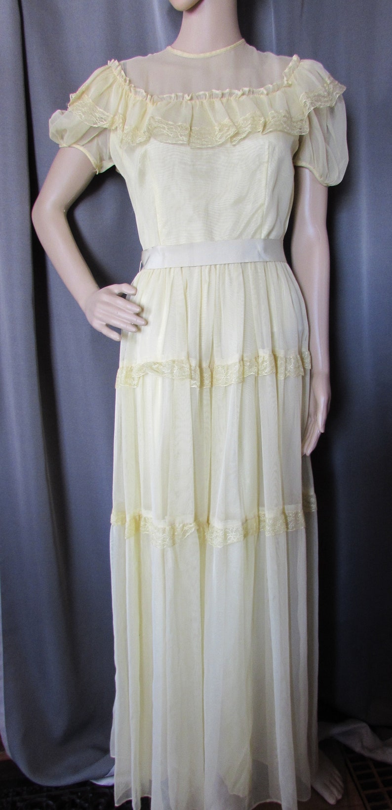 May include: A pale yellow, tiered dress with a lace trim and a satin sash. The dress has a sheer, ruffled neckline and short sleeves.