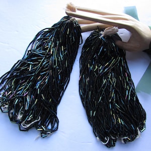 Pode incluir: Duas meadas de contas de vidro preto iridescente, cada meada é agrupada e amarrada com um cordão branco. As contas são longas e estreitas, parecidas com pequenos tubos.