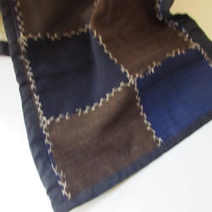 May include: A patchwork textile with squares of brown and navy blue fabric, stitched together with white thread. The edges are finished with a dark blue border. The textile is folded, revealing a portion of the back.