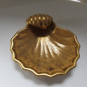 Puede incluir: Un plato de cerámica en forma de concha de color dorado con un borde acanalado y un asa de concha elevada. El plato tiene un acabado texturizado y metálico.
