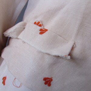 May include: Close-up of a cream-colored fabric with orange embroidered floral designs. The fabric has a slightly textured surface.