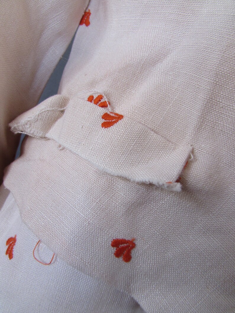 May include: Close-up of a cream-colored fabric with orange embroidered floral designs. The fabric has a slightly textured surface.